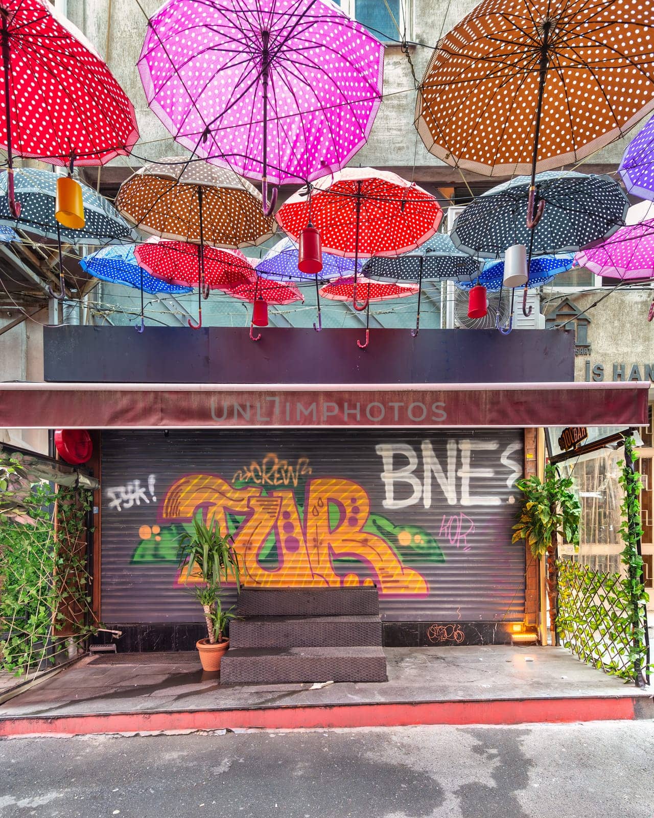 Istanbul, Turkey - May 13, 2023: Group of colorful umbrellas hanging over a closed shop rolling door with colorful graffiti in a narrow alley in Karakoy neighborhood