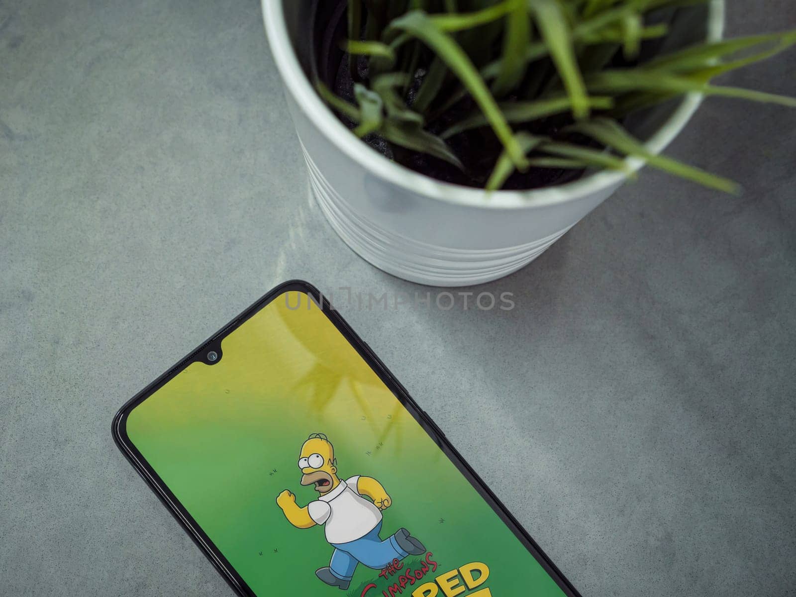 Lod, Israel - July 16,2023: Modern workspace with smartphone with The Simpsons Tapped Out app launch screen on marble background. Close up top view flat lay.