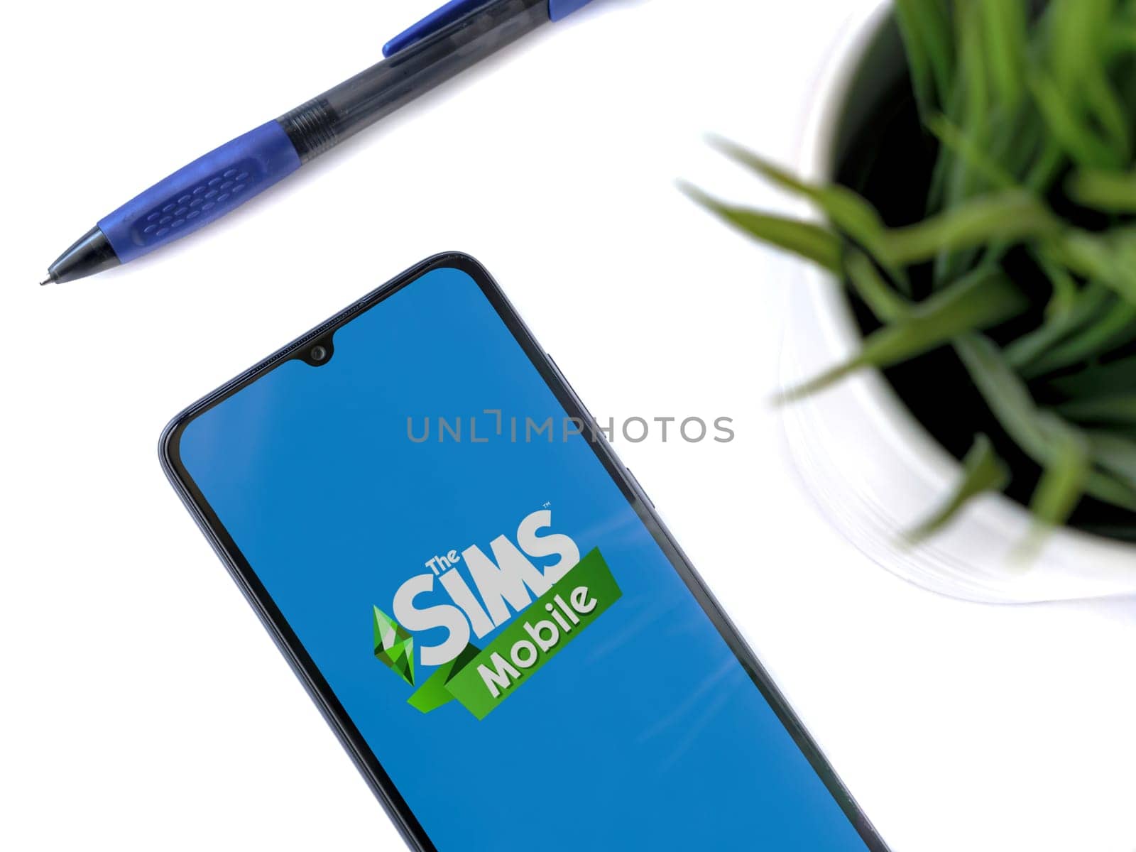 Lod, Israel - July 16,2023: Modern workspace with smartphone with The Sims Mobile app launch screen on white background. Close up top view flat lay.