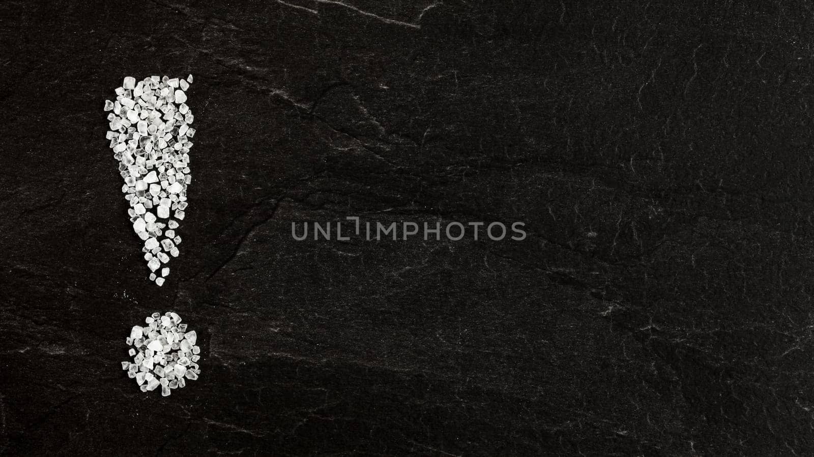 Exclamation mark made of sea salt crystals on black slate board. Wide banner with space for text (unhealthy oversalt concept) right.