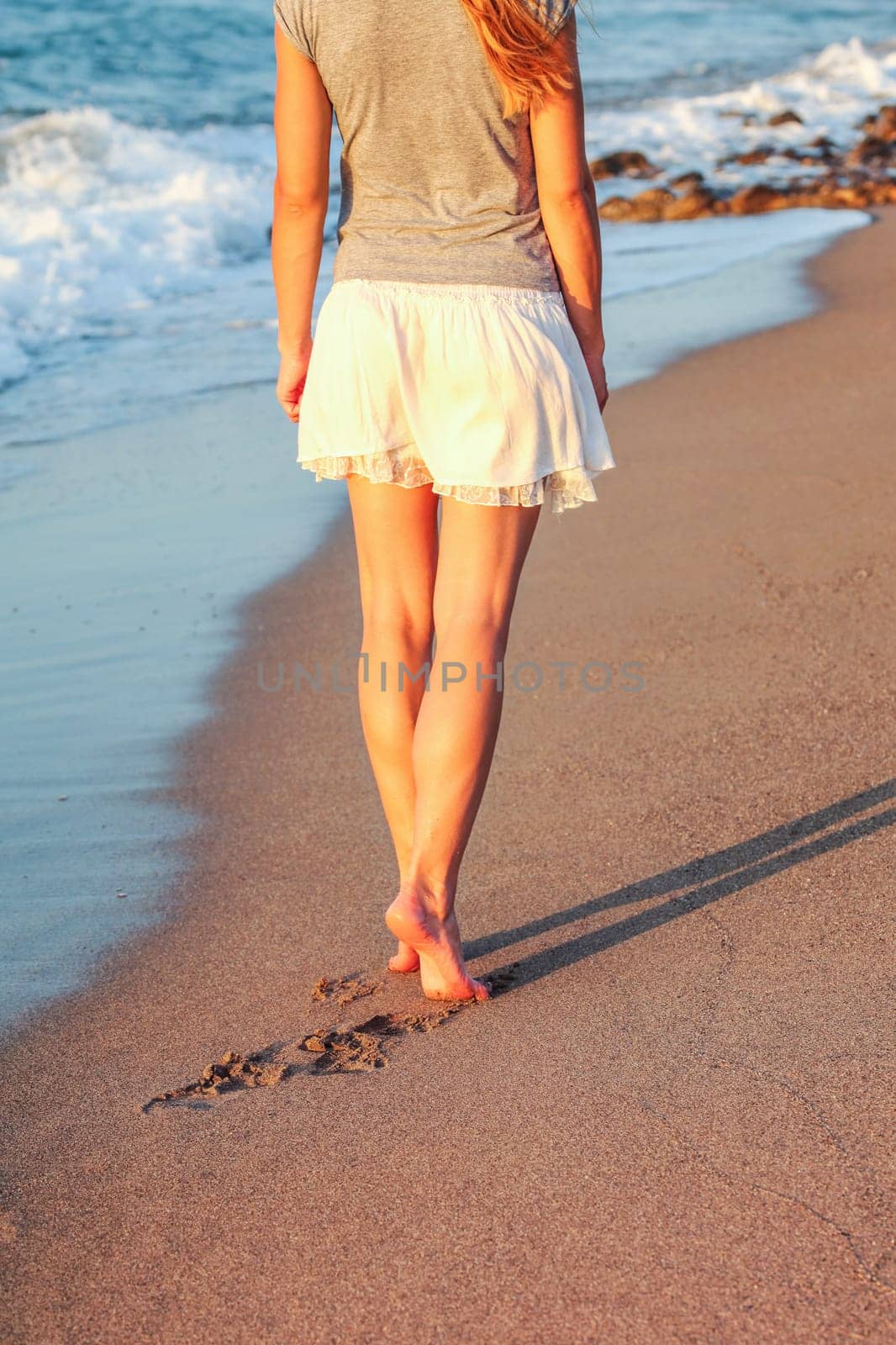 Young woman walking on wet sand beach lit by sunset light, view from back, detail on her calf leg muscles