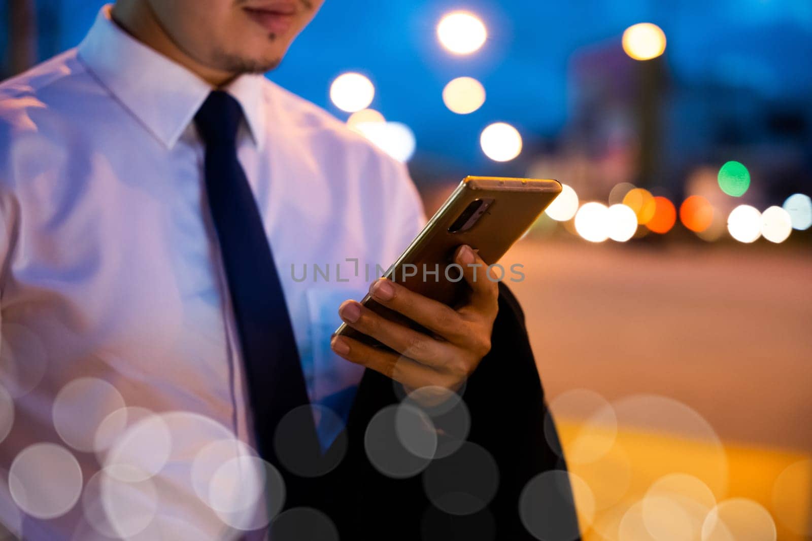 Portrait Asian businessman typing an sms message via smartphone after work near office at night city street, bokeh lights, young man walking and chatting with friends at social networks outdoor