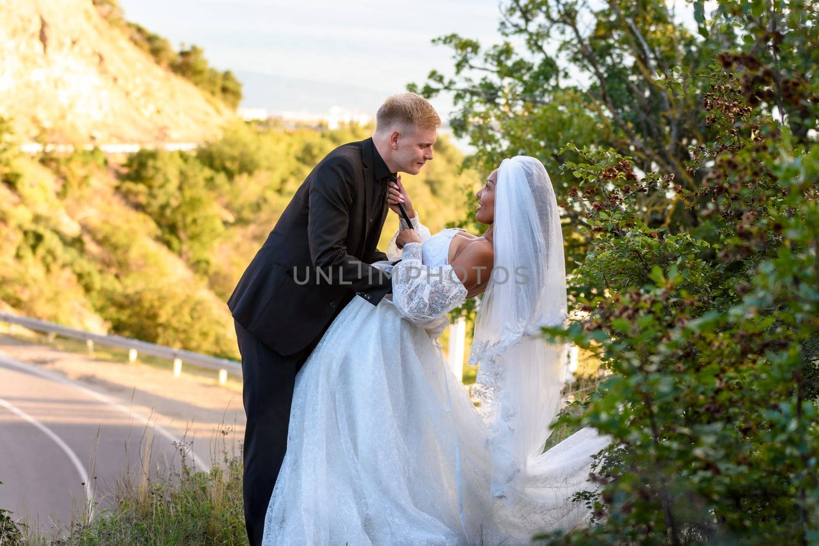 Happy newlyweds kiss against the backdrop of a beautiful mountain landscape, the girl holds the guy by the tie