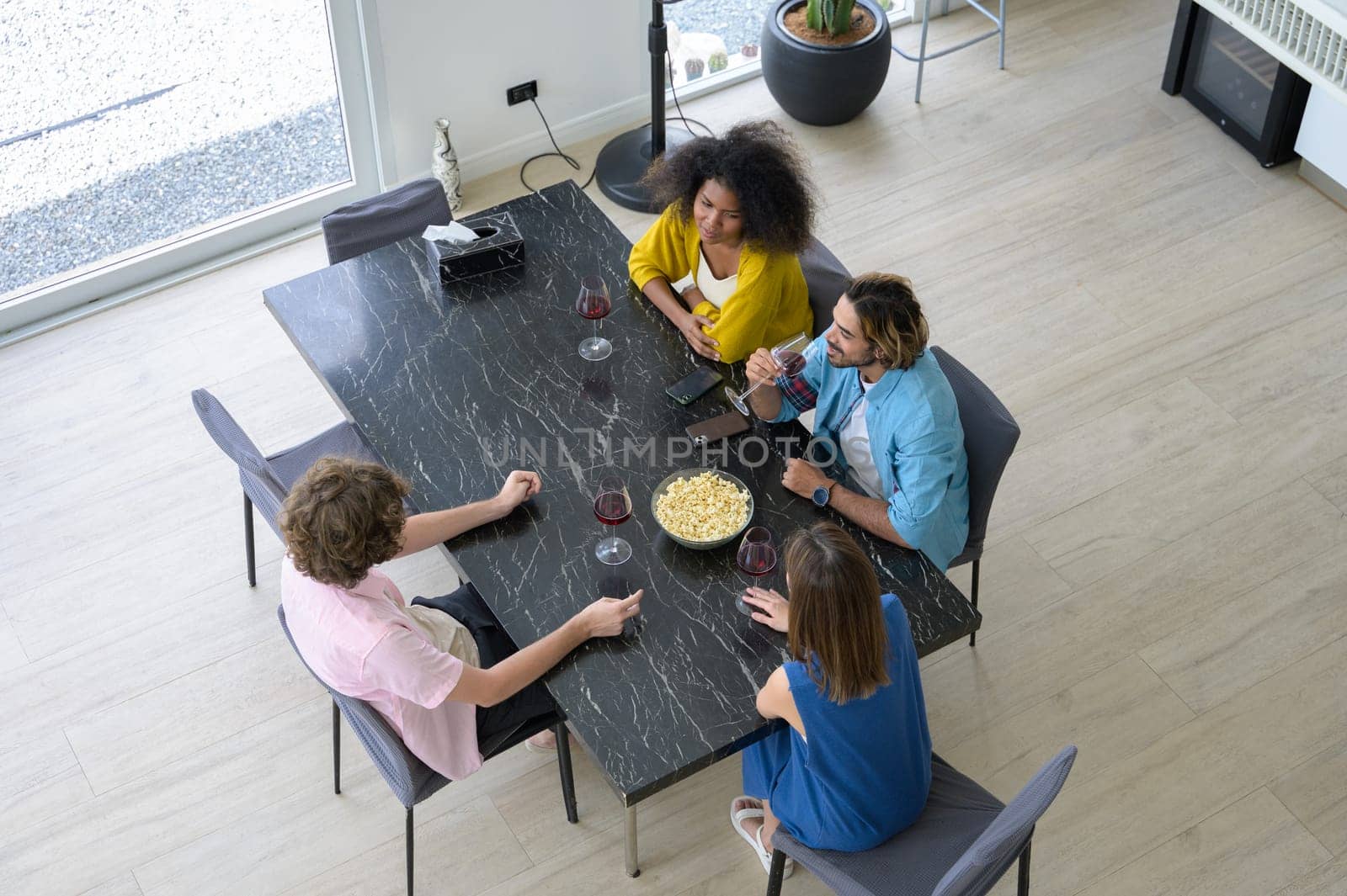 Multiracial group of happy young people sitting around table, drinking red wine with popcorn and having conversation together at home.