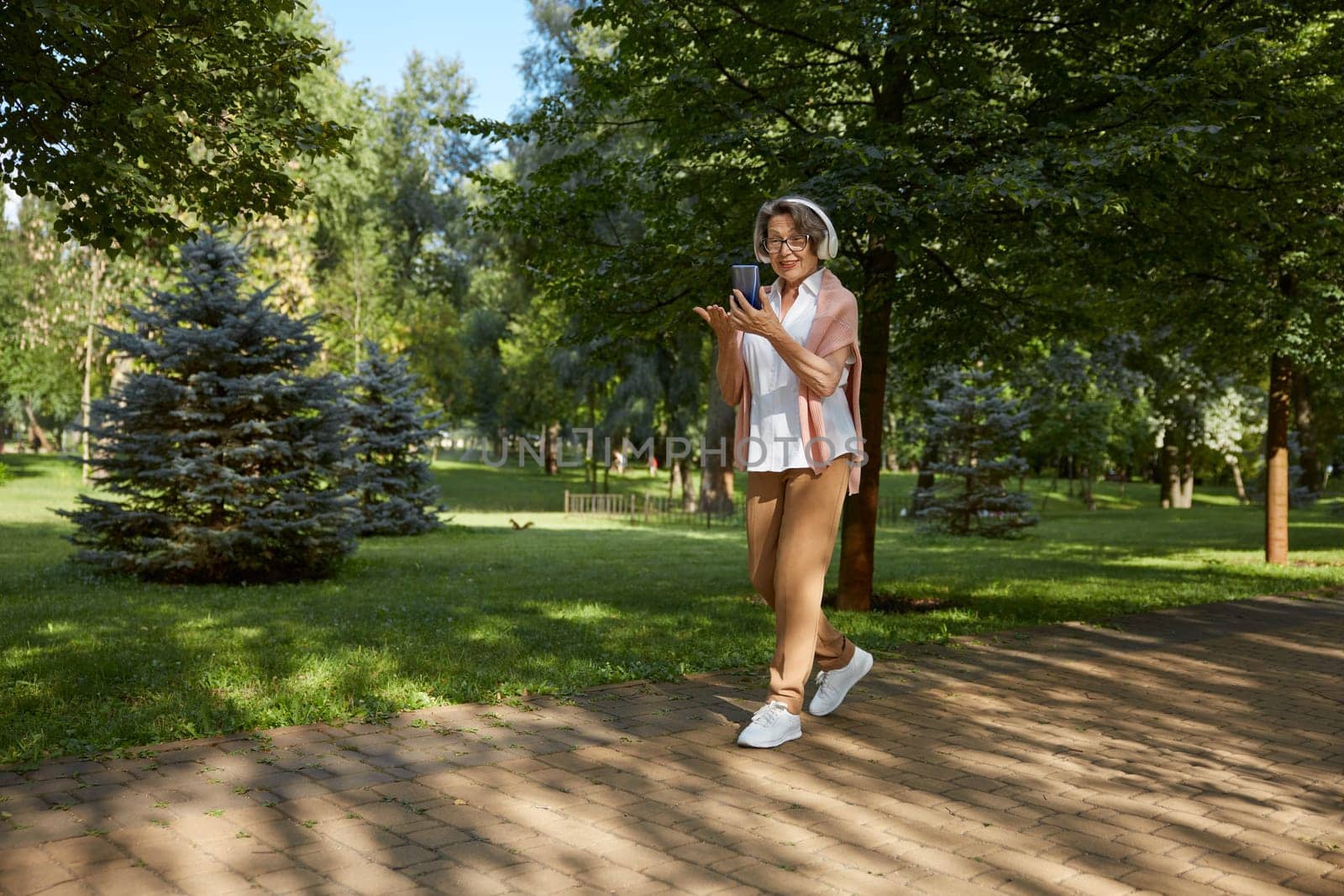 Elderly old woman in headset having online video call on smartphone using wireless internet connection with relatives or friends in city park