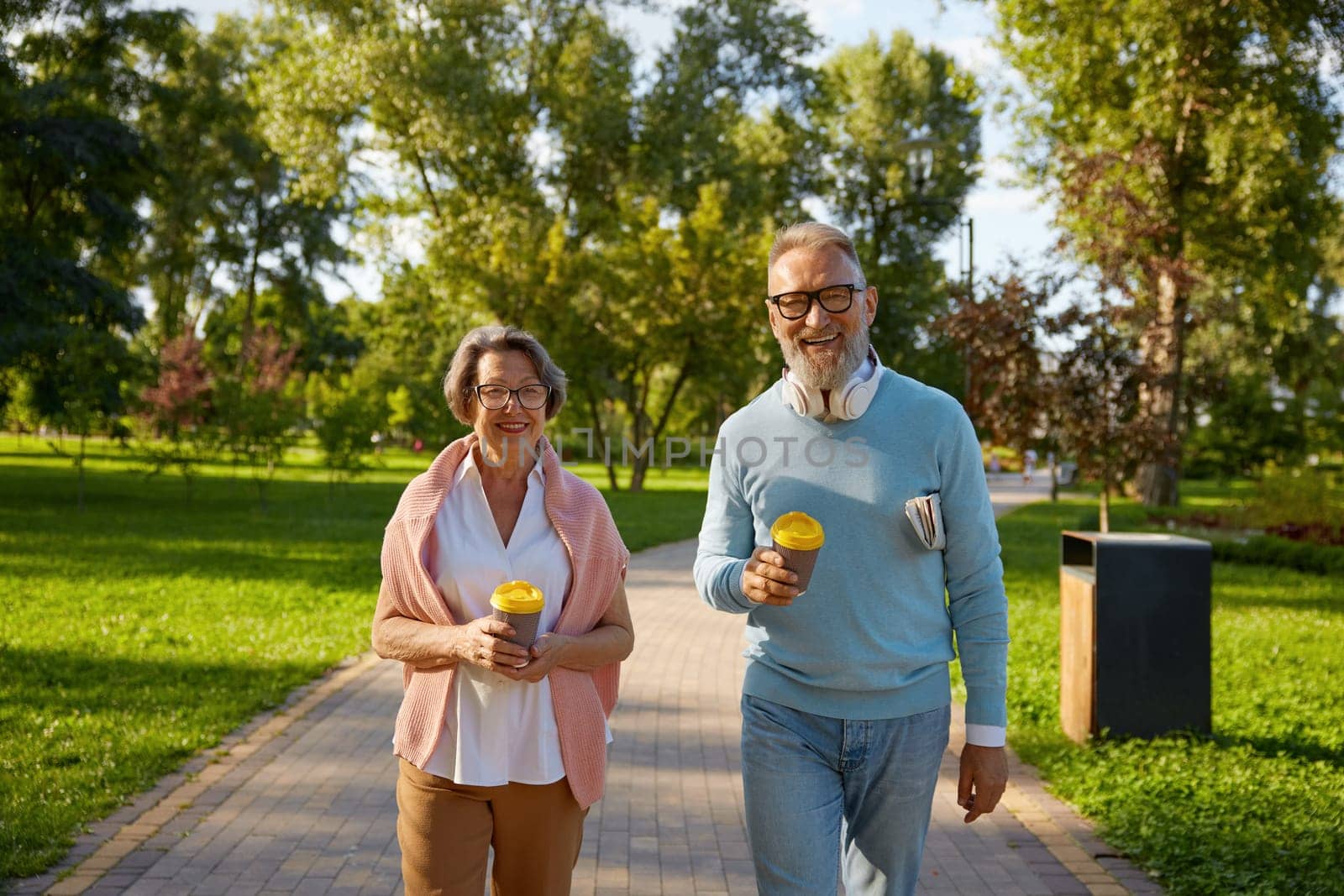 Happy fashioned elderly couple having coffee break walking in park. Romantic aged mature man and woman looking happy enjoying healthy lifestyle on pension