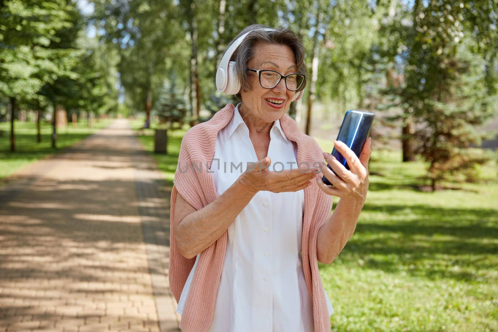 Elderly old woman in headset having online video call on smartphone using wireless internet connection with relatives or friends while walking in park