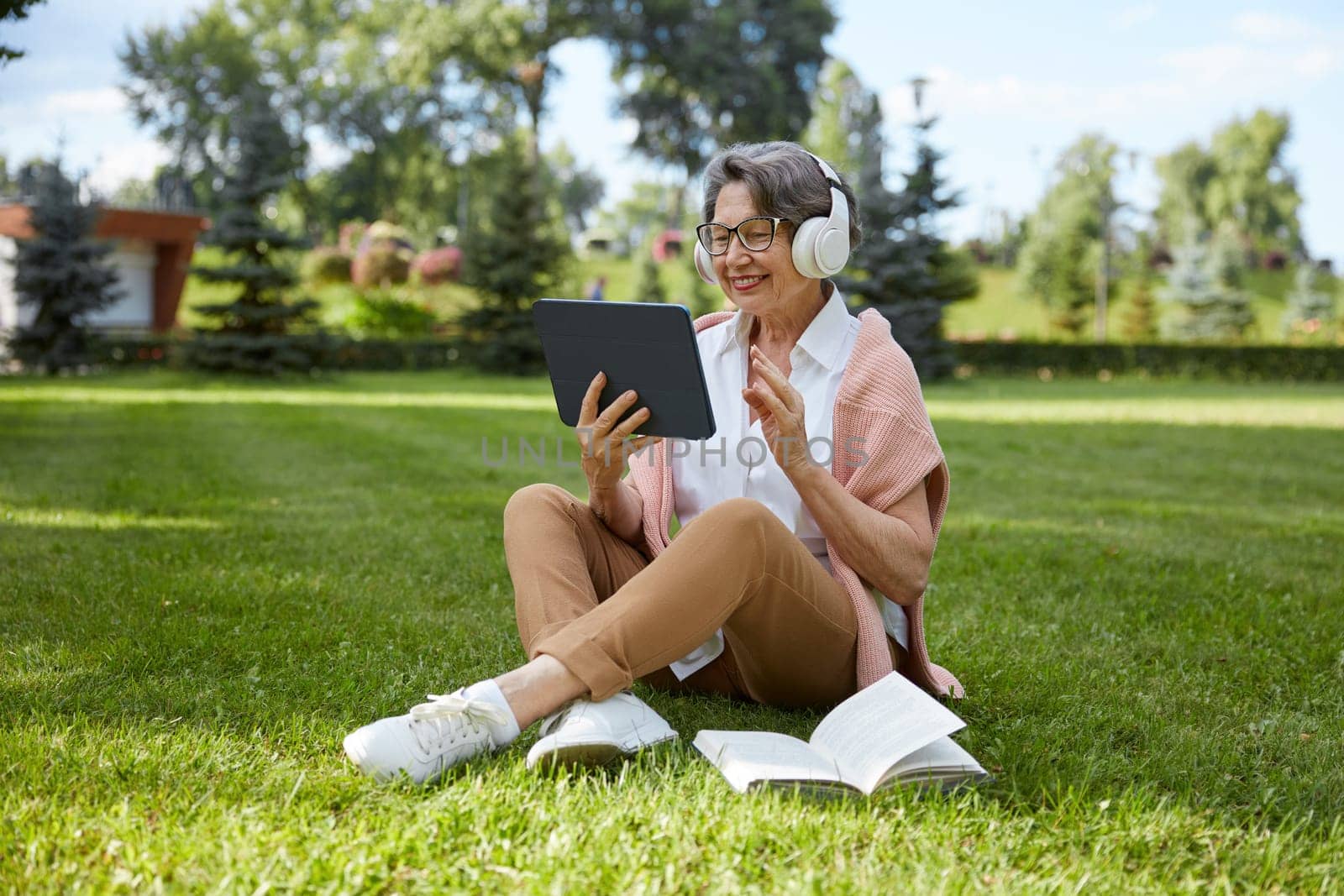 Happy smiling elderly woman talking with friends or having education webinar with teacher using videocall app on mobile digital tablet while resting in public park
