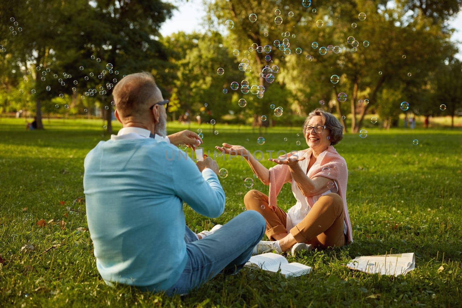 Playful active senior couple blowing soap bubbles while rest in park. Vitality and active elderly people concept