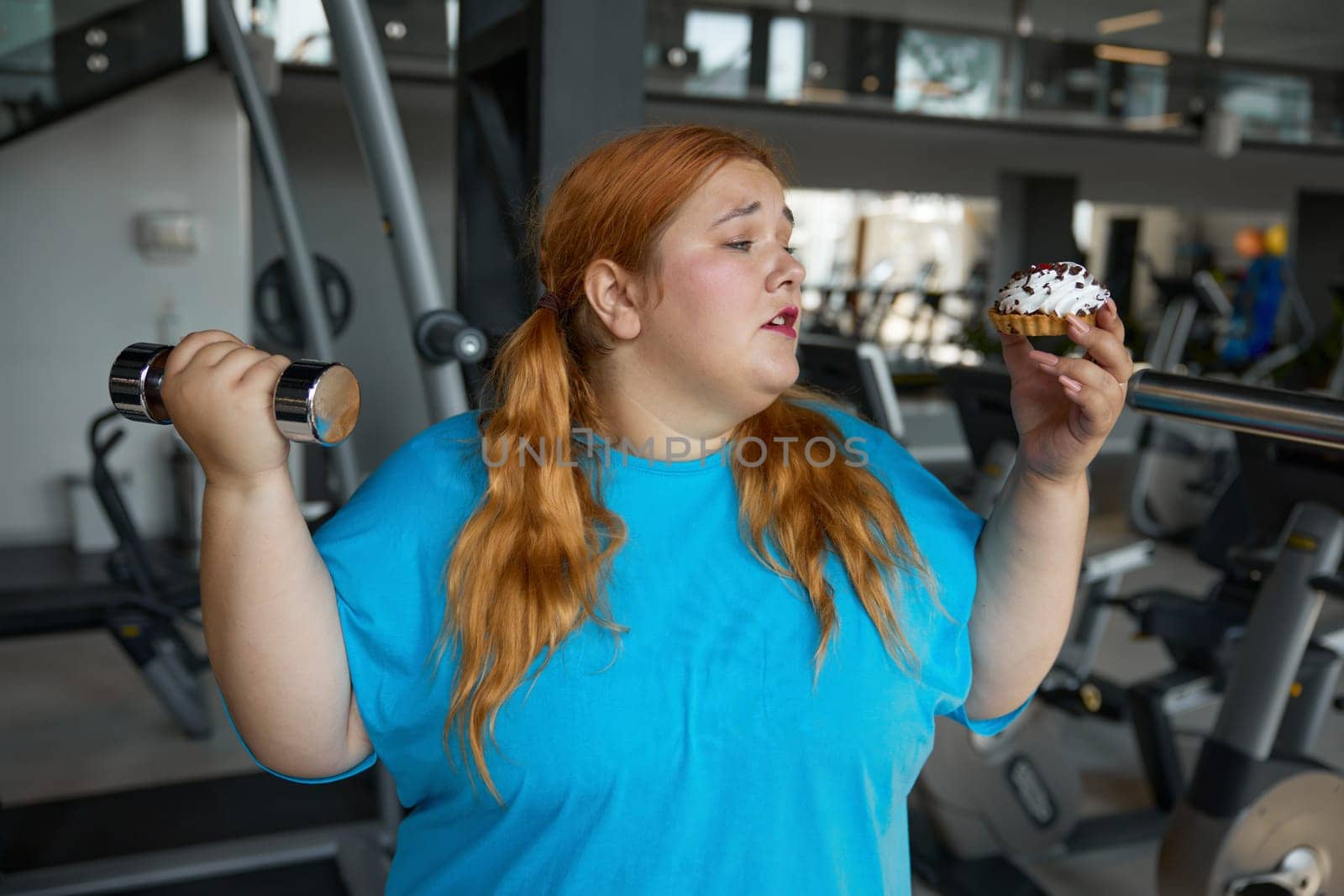 Portrait of comic overweight woman holding dumbbell looking with irresistible desire at donut. Sport and fitness workout for slimming