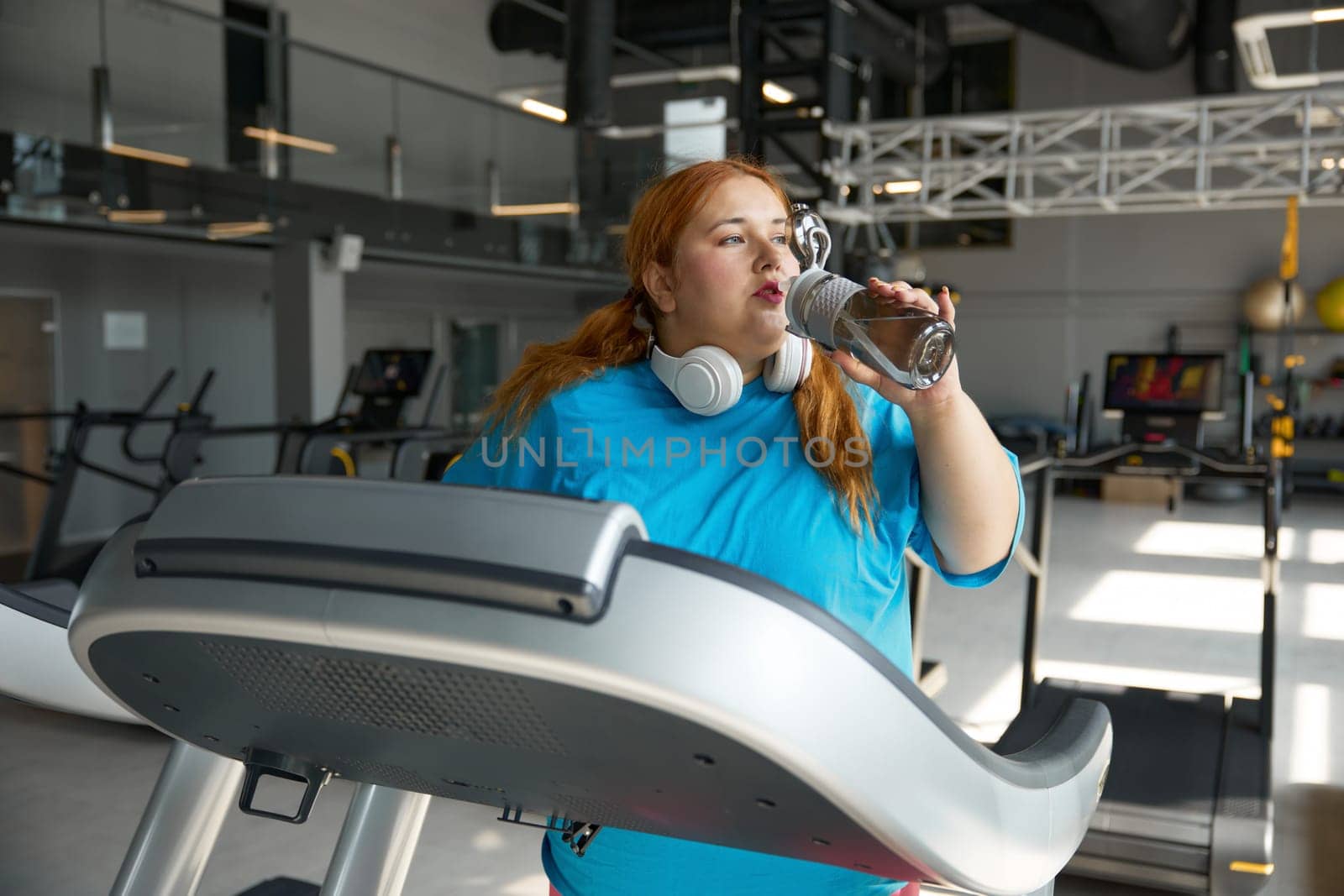 Exhausted sportive overweight woman drinking water taking break during training on treadmill machine. Cardio workout at professional gym
