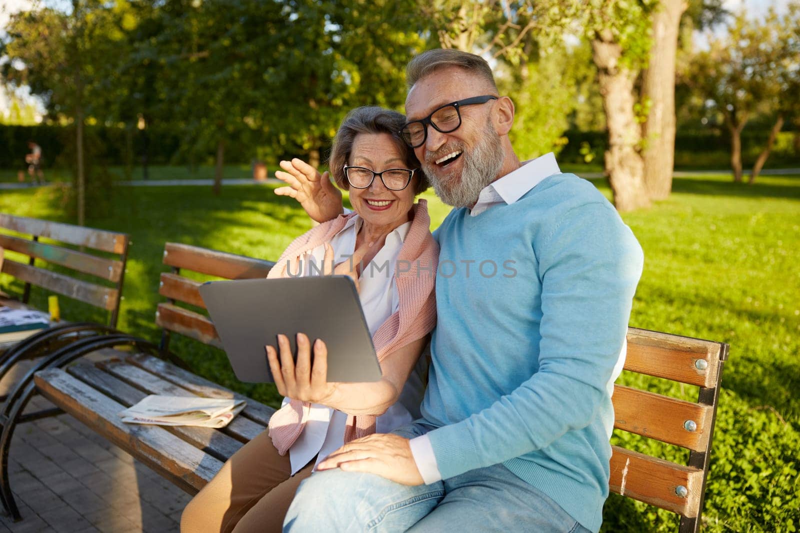 Smiling senior couple in stylish clothes having videocall conversation using digital tablet sitting on park bench. Elderly people and technology