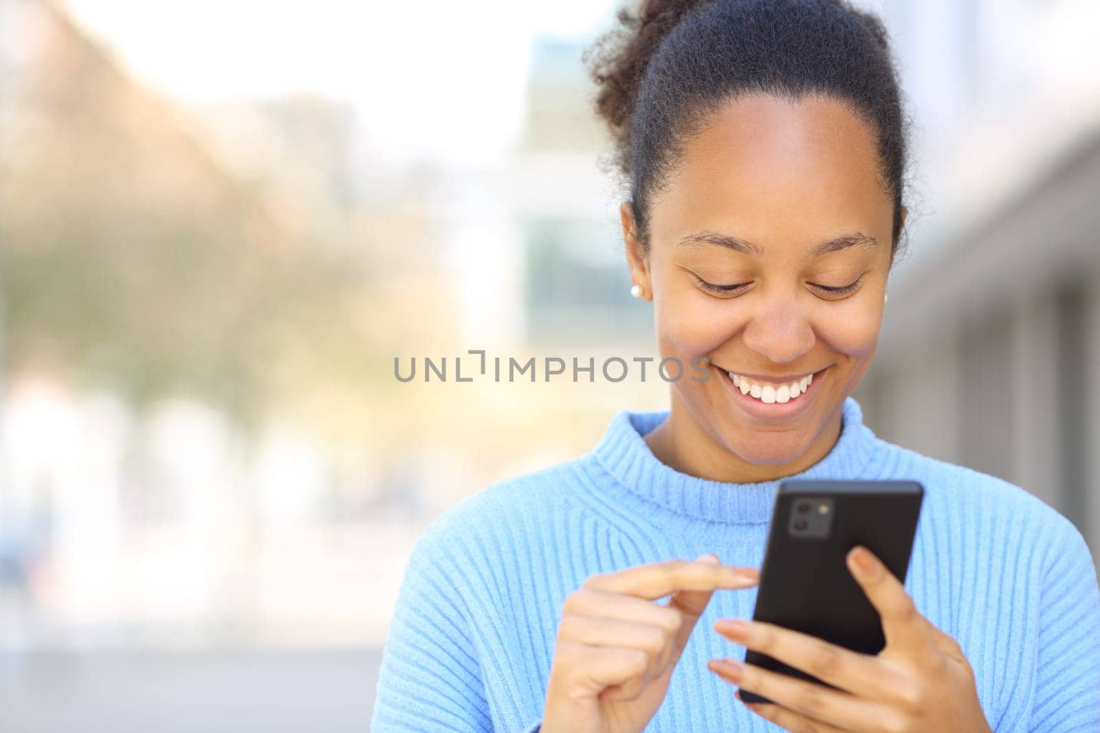 Front view portrait of a happy black woman using cell phone in the street with copy space