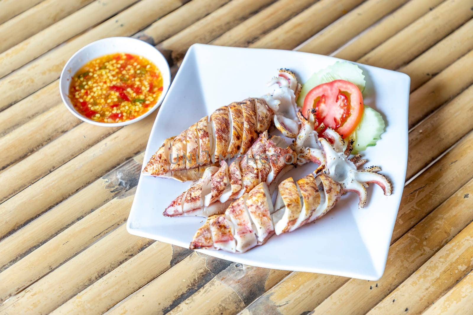 3 grilled squid sit on clean white plate on bamboo table alongside slices of tomato and cucumber. There is a bowl of spicy seafood dipping sauce next to it. It's fresh seafood in a seaside restaurant.