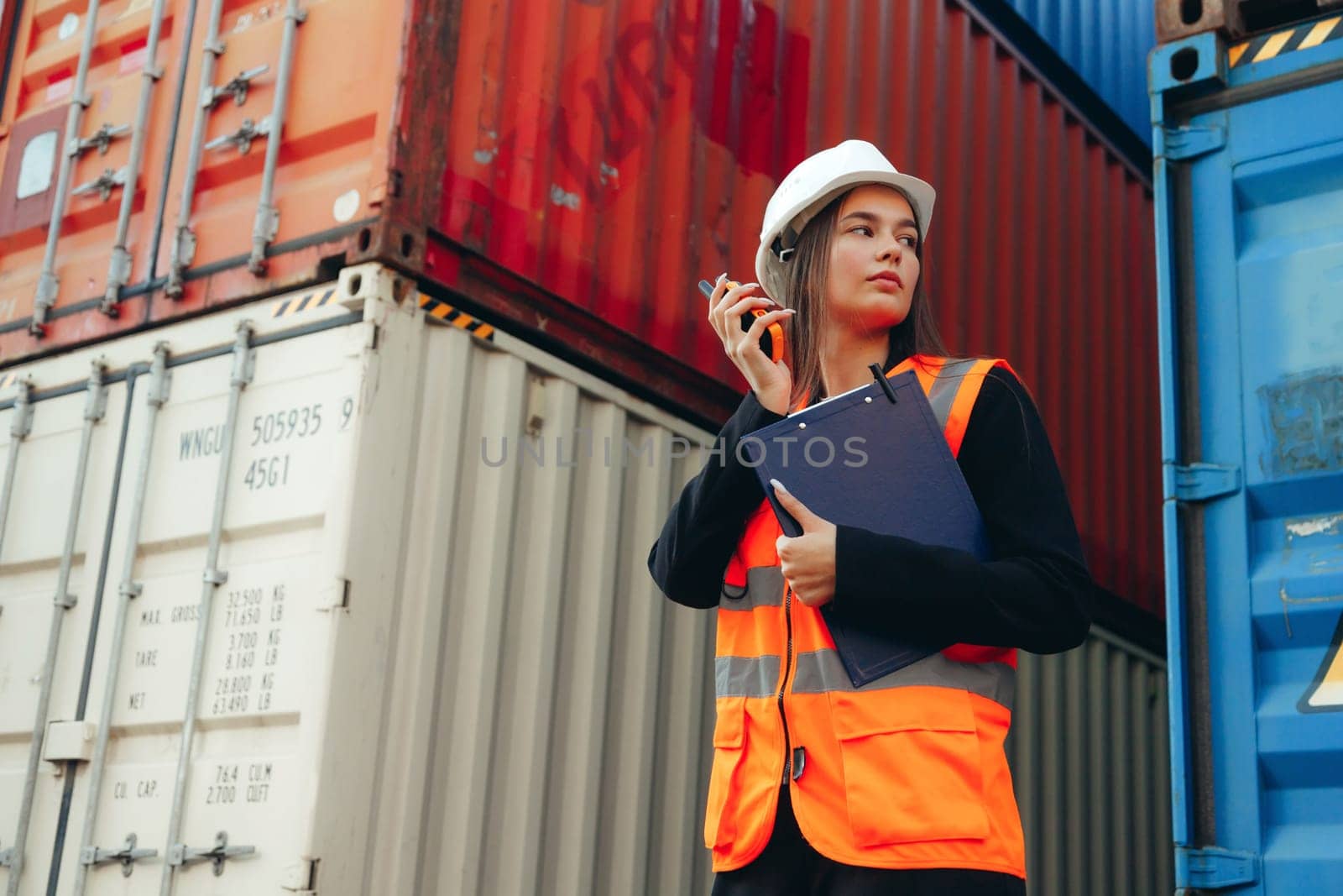 Portrait of beautiful engineer woman with walkie-talkie in white helmet and vest working in container terminal port.