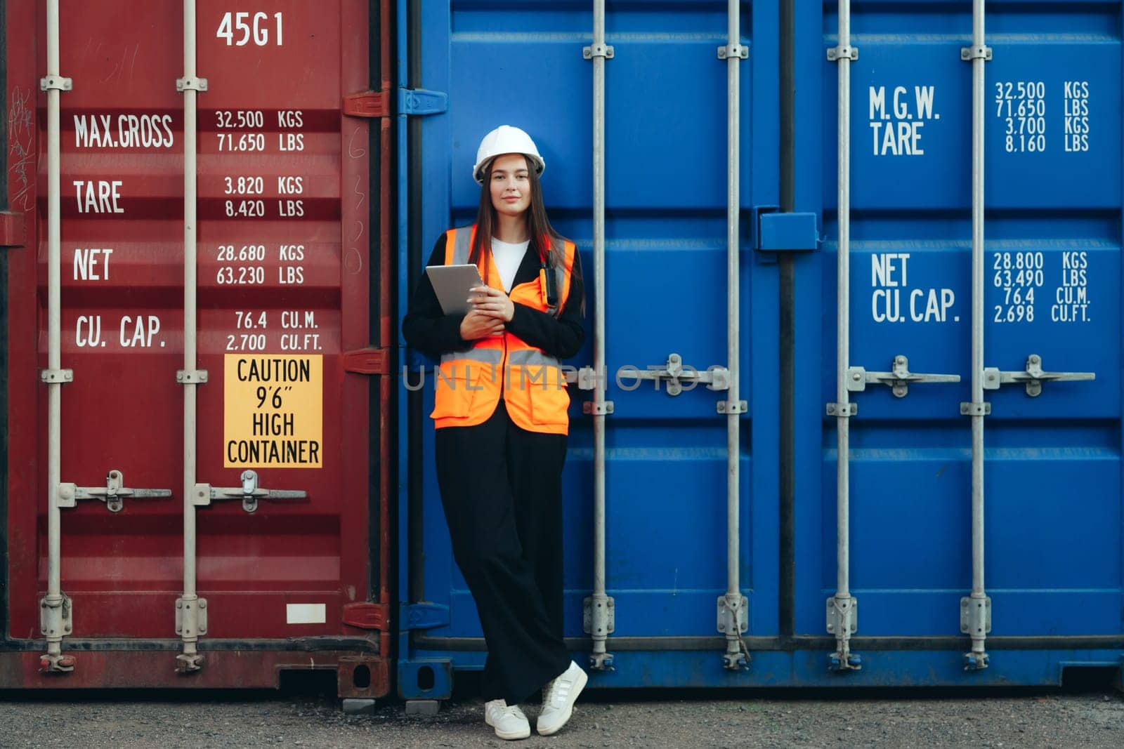 Female Engineer Containers Terminal Worker with tablet posing at container. White helmet and vest in container terminal port or construction site.