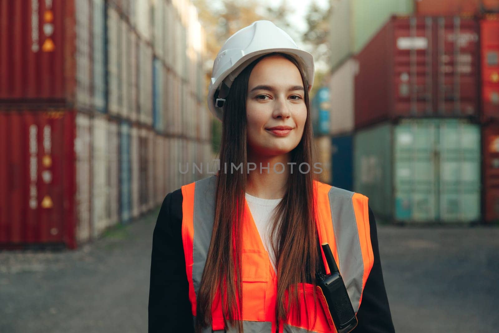 Portrait of beautiful engineer woman with walkie-talkie in white helmet and vest working in container terminal port.