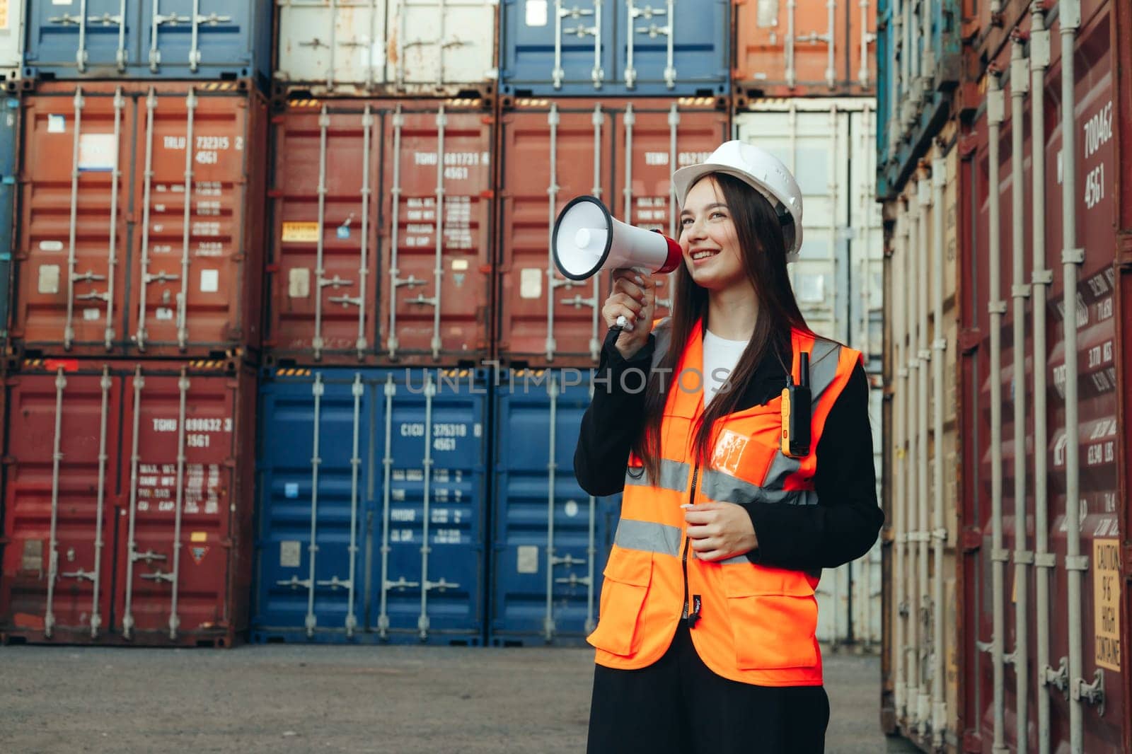 Female engineer speaking into a megaphone while standing in an industrial warehouse or terminal with shipping containers.