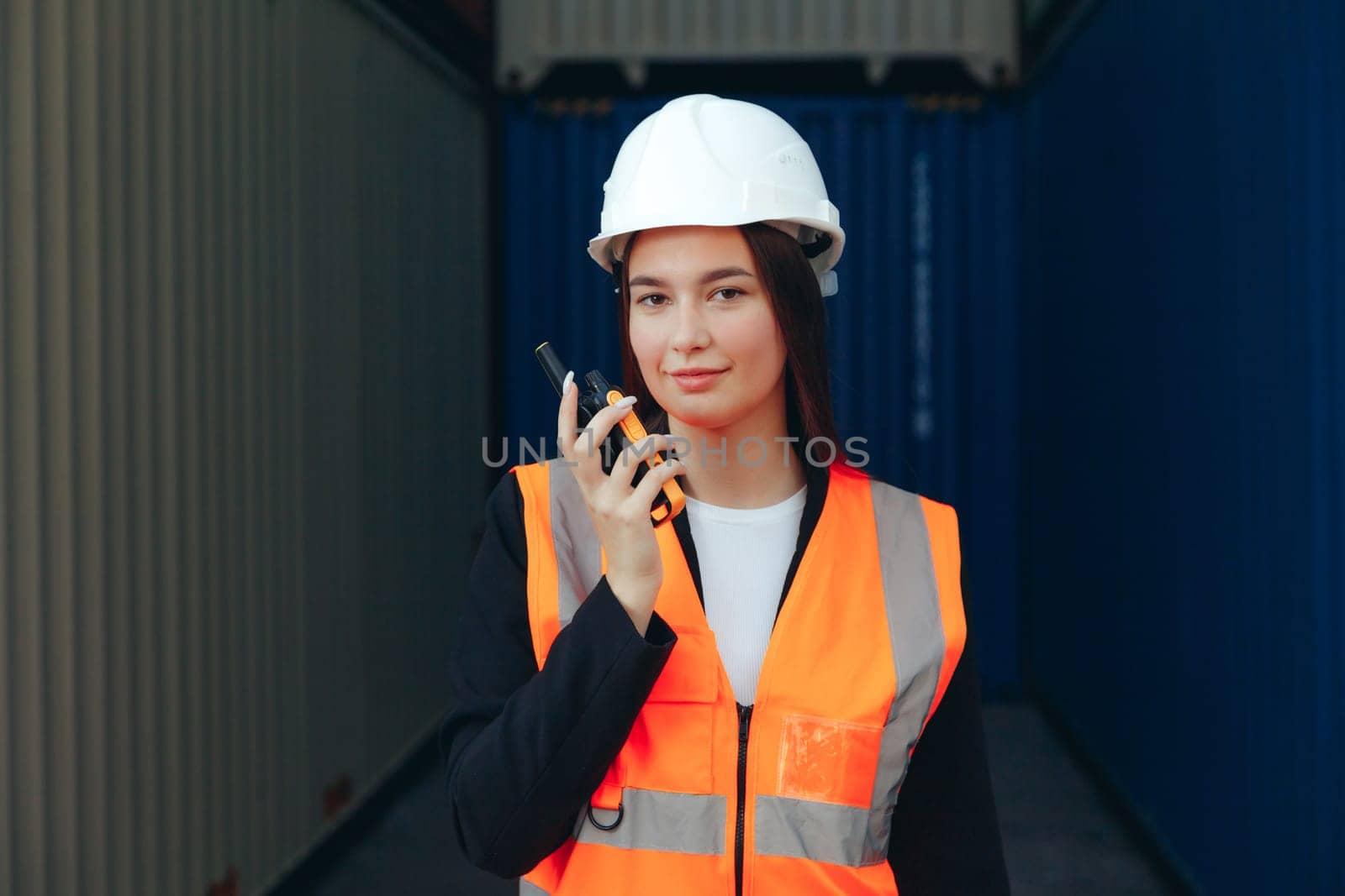 Portrait of beautiful engineer woman with walkie-talkie in white helmet and vest working in container terminal port.