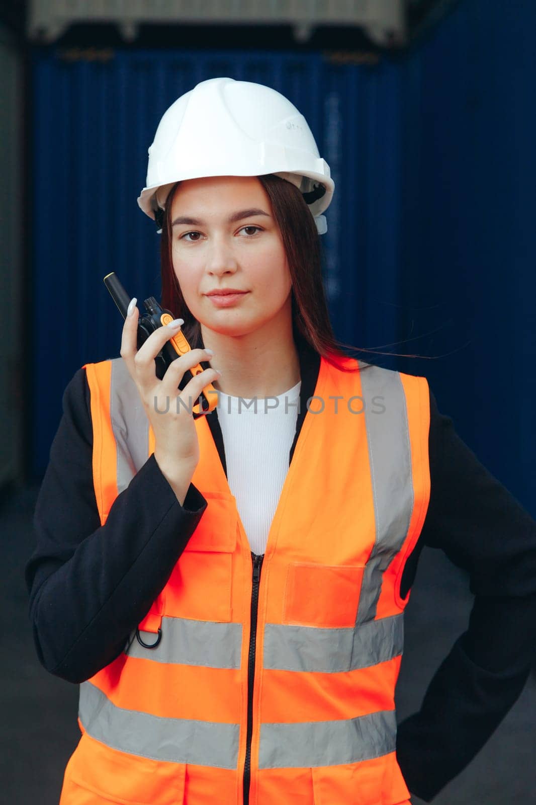 Portrait of beautiful engineer woman with walkie-talkie in white helmet and vest working in container terminal port.