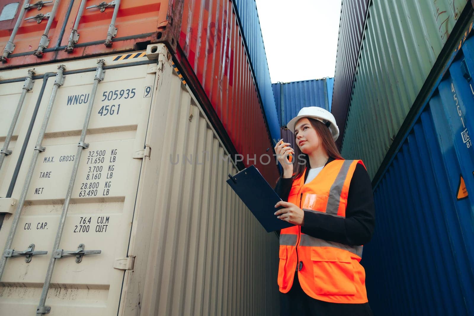 Female Foreman using walkie talkie radio and clip folder, control loading containers box. Engineer or worker with safety hat checking industrial container cargo freight ship