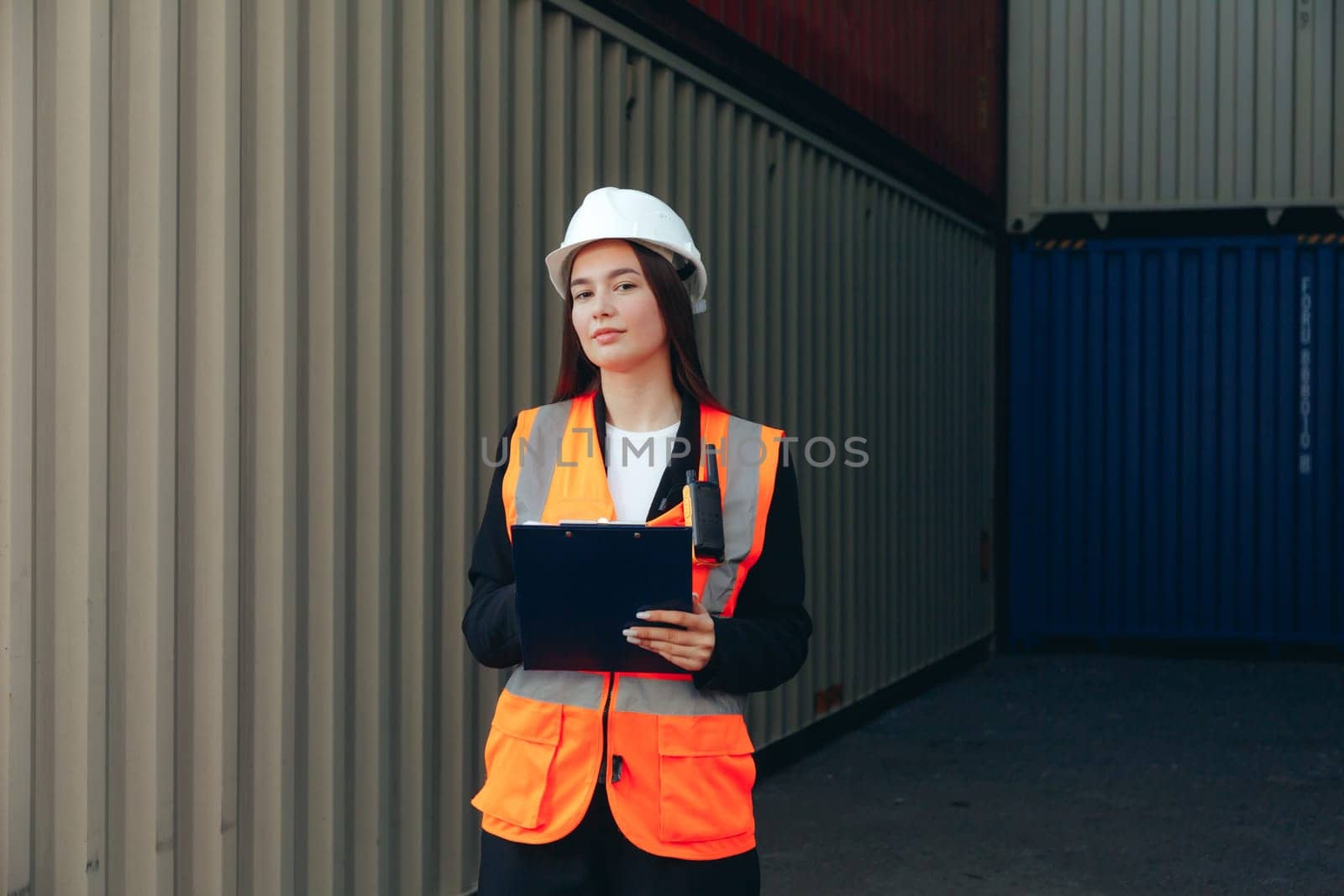 Female Engineer with Clip Folder in white helmet and vest working in container terminal port or construction site.