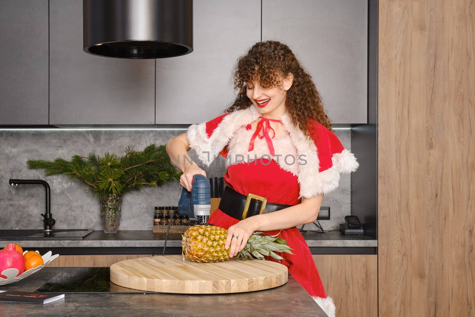 Laughing woman in christmas costume cutting pineapple with jigsaw in the kitchen