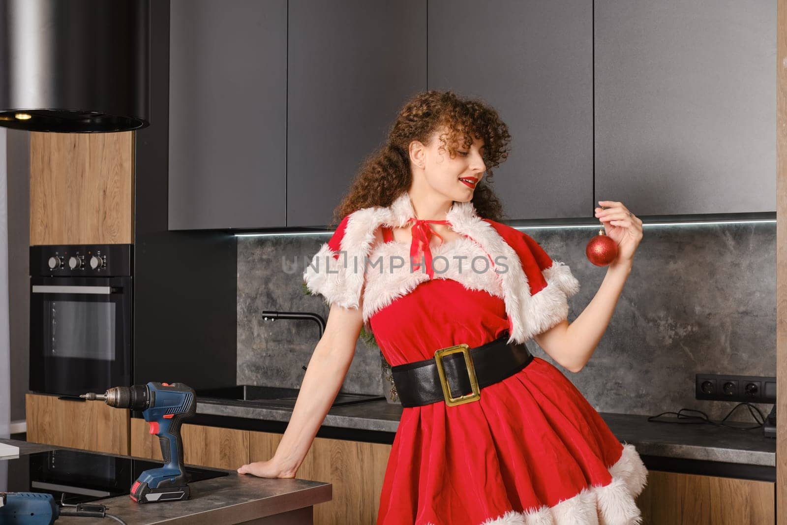Christmas gift concept - new kitchen furniture. Cheerful woman in santa dress looks at toy ball.