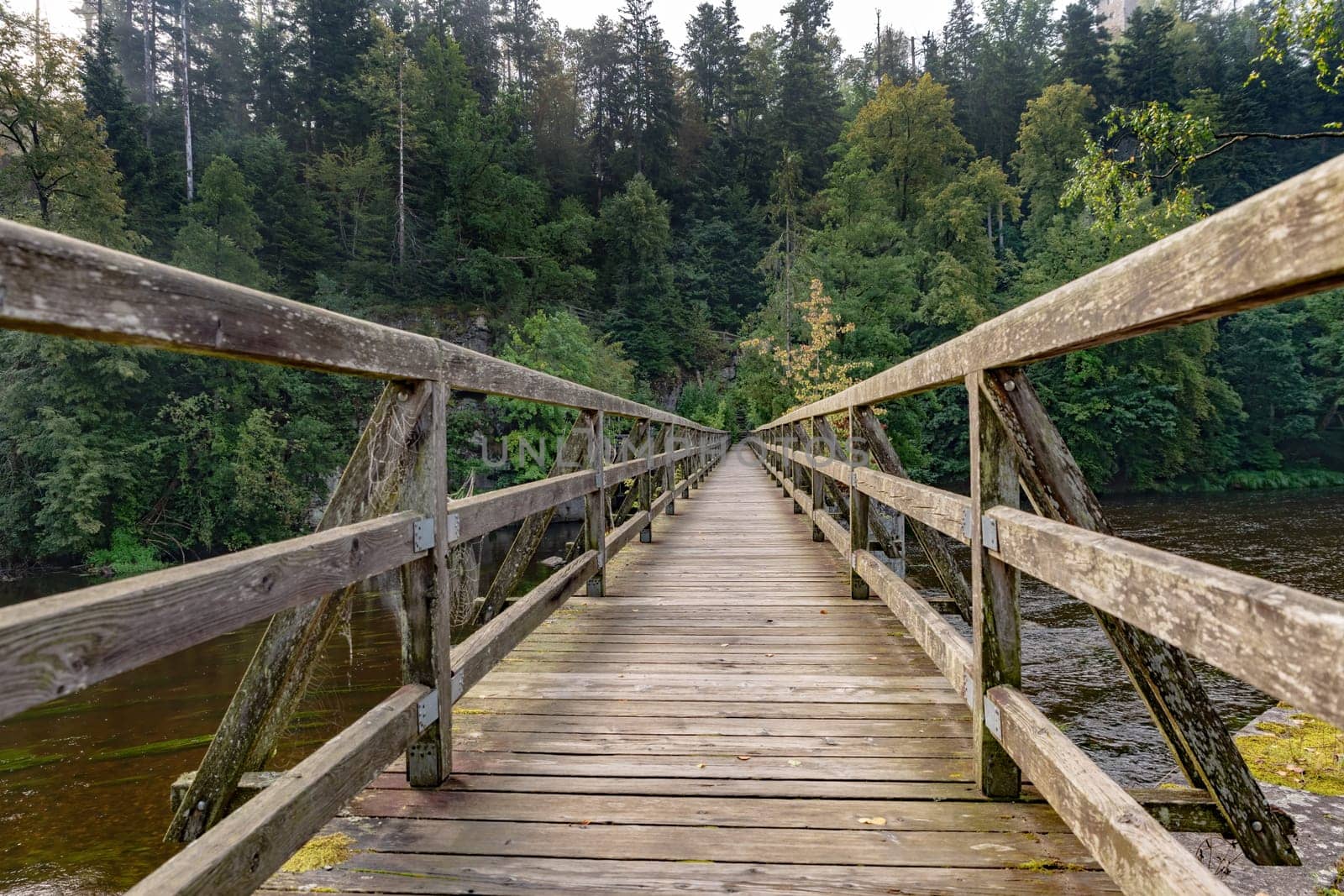 Bridge made of wooden boards over a green field with trees and wooded hills
