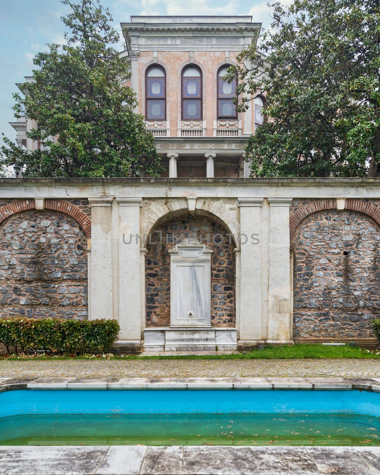 A beautiful white marble drinking fountain located in the garden of the Mecidiye Pavilion, Istanbul, Turkey, with a decorative fountain in front, and the palace in the background