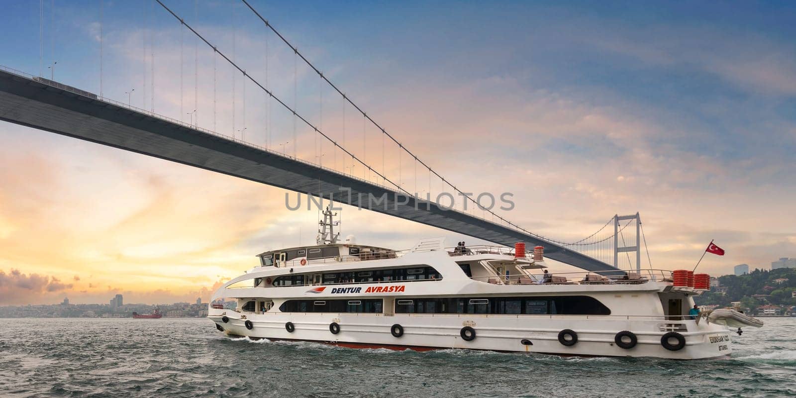 Istanbul, Turkey - May 17 2023: White ferryboat sails under the Bosphorus Suspension Bridge before sunset, with the city skyline in the background. Photo taken from Beylerbeyi Palace