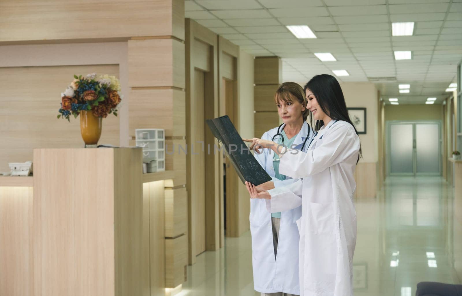 Two women doctor working together examining patient x-ray scan in hospital . Senior doctor and young smiling doctor standing in hallway and discussing diagnosis of brain x ray results. Healthcare, medicine and insurance concept