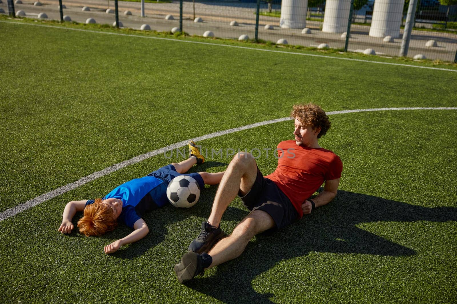 Tired father and son soccer player looking sweaty and exhausted lying on grass of football field. Family sport leisure concept