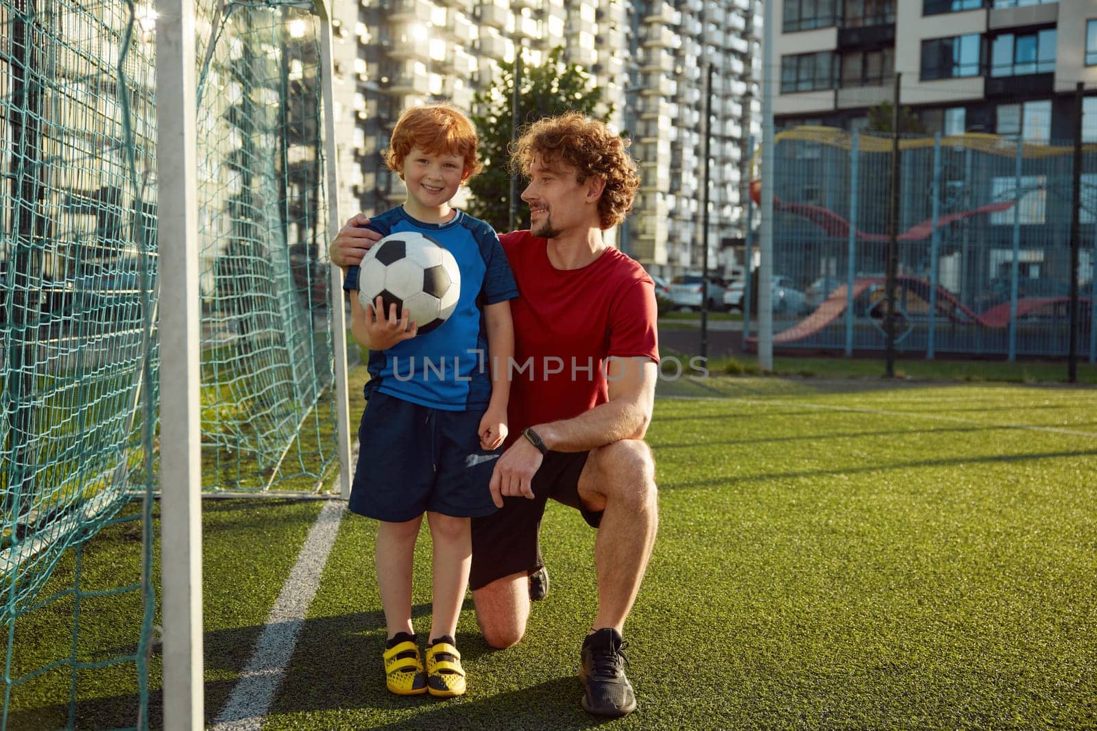 Portrait of happy father and little son standing near football gate. Family playing soccer on outdoor field