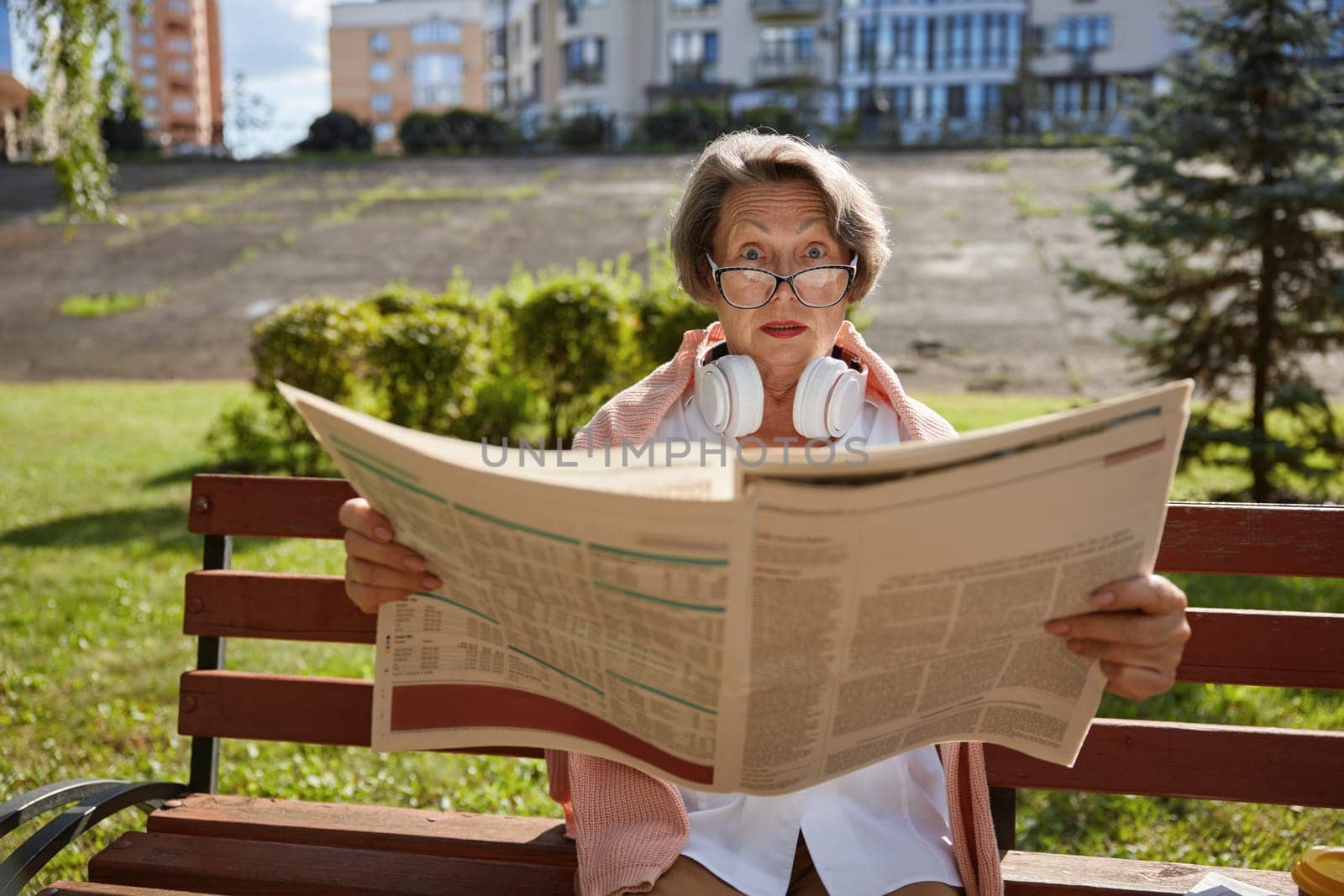 Surprised or shocked elderly woman reading daily newspaper finding overwhelmed news in morning in public park