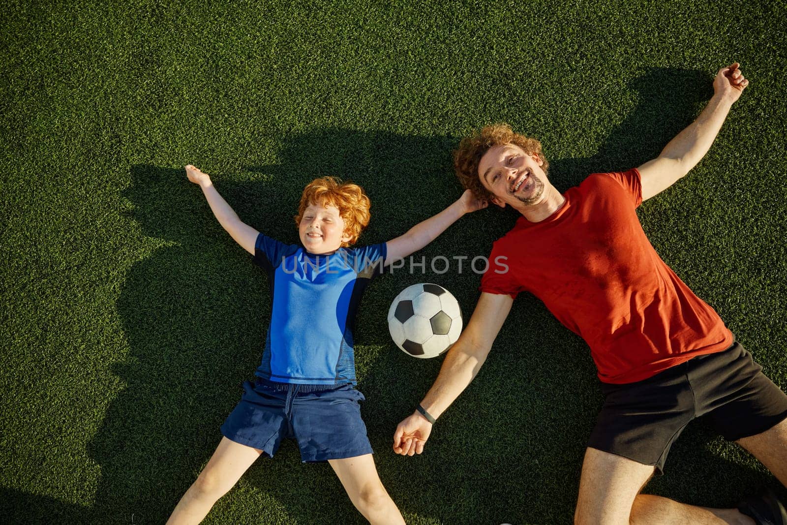 Overjoyed father and little son child smiling having fun while lying on soccer field grass overhead view. Happy family sport recreation