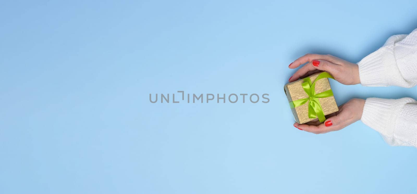Female hands holding a gift box with a green ribbon on a blue background. View from above, copy space