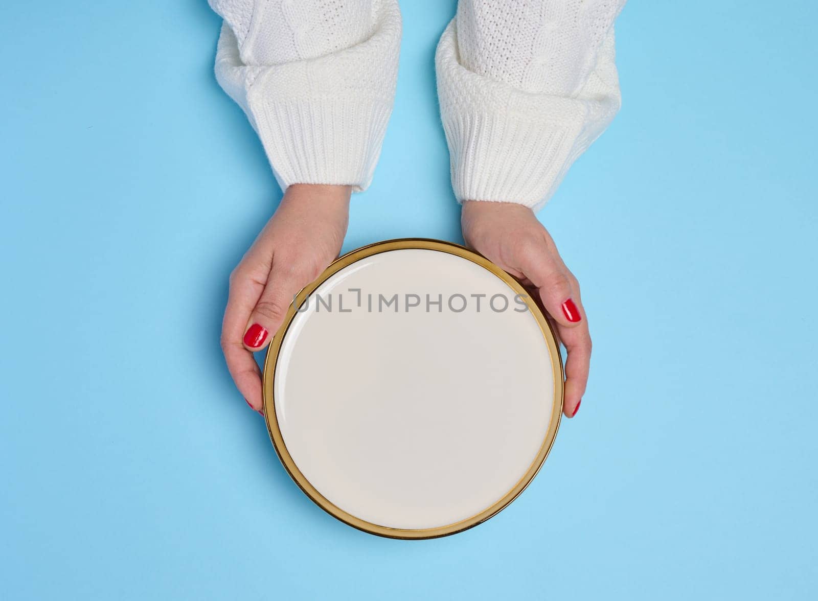 Two female hands holding an empty round white ceramic plate on a blue background, top view
