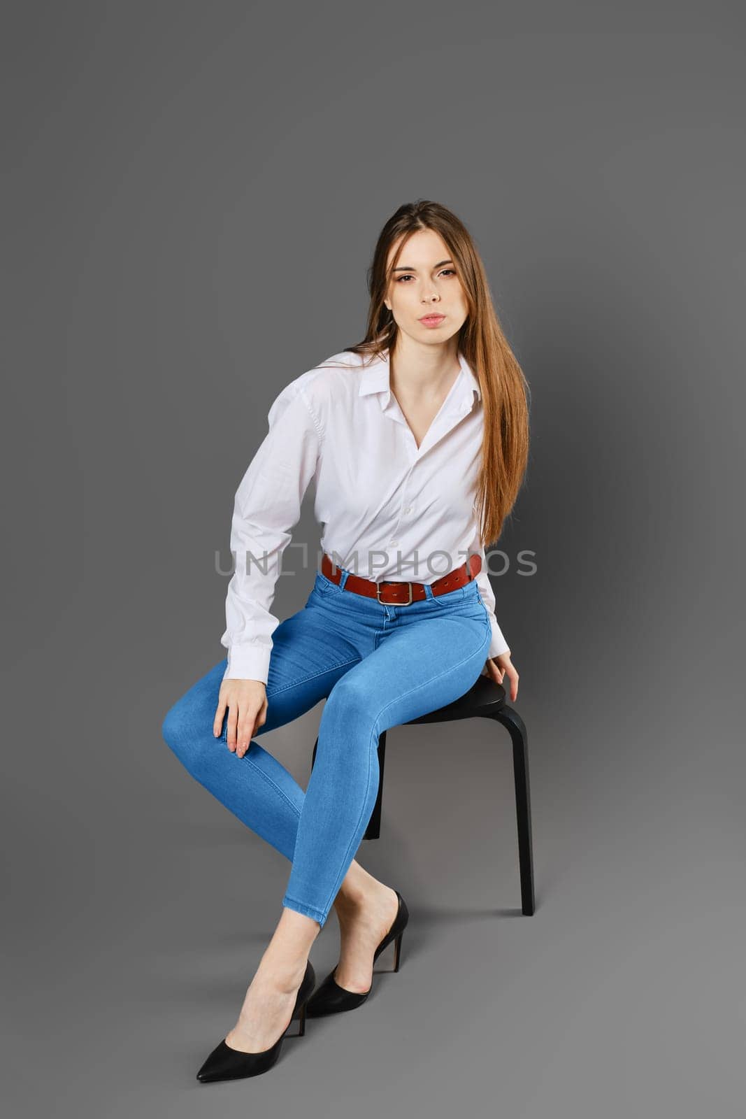 A young woman in a white shirt and blue jeans sits on a chair in a tense pose on a grey studio background