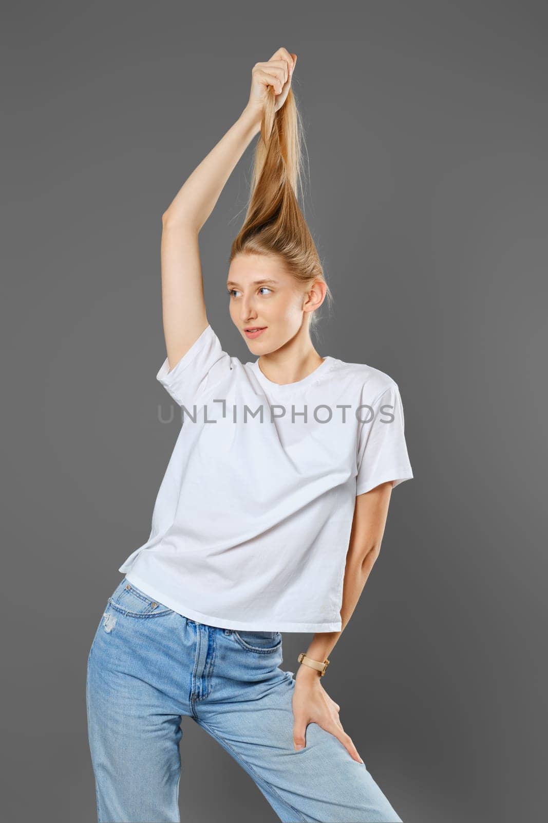 Playful woman wearing blue jeans and white shirt playing with her long blonde hair in studio over grey background