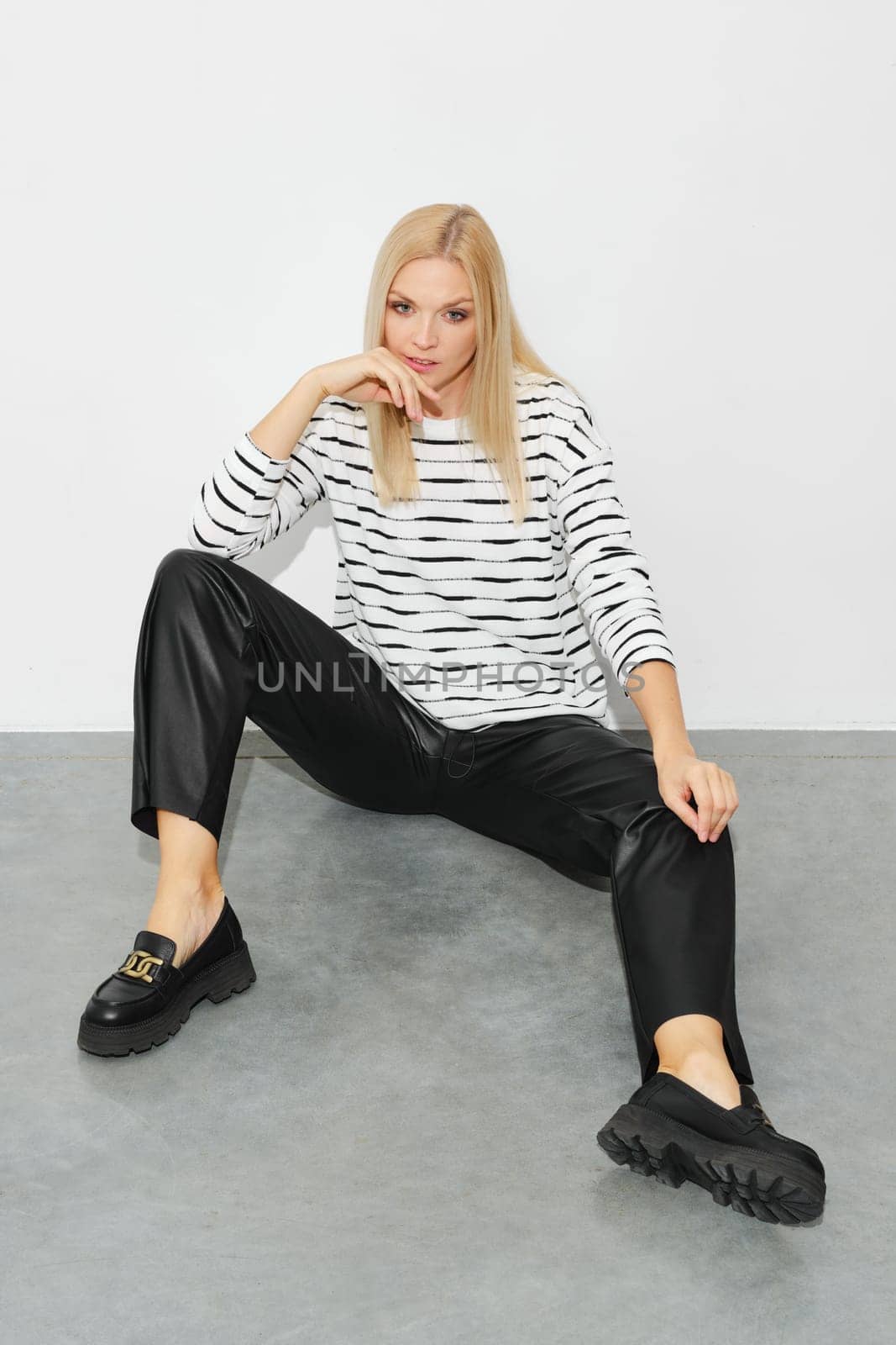 Stylish relaxed woman in a striped sweater and leather pants sits on the floor