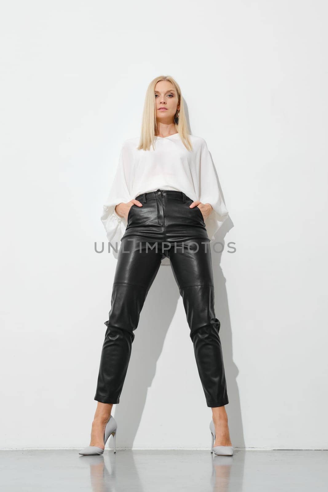 Low angle view at blonde woman in black leather trousers and white blouse leaning against white wall
