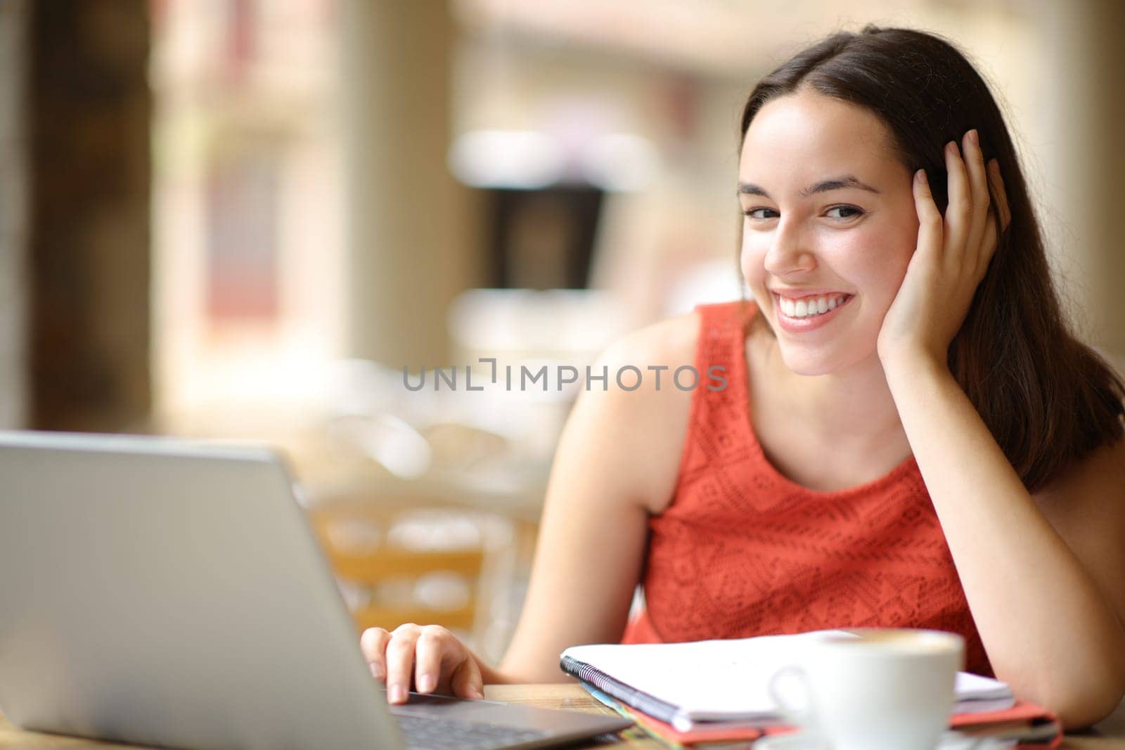 Happy student with notebook and laptop looks at you in a coffee shop terrace