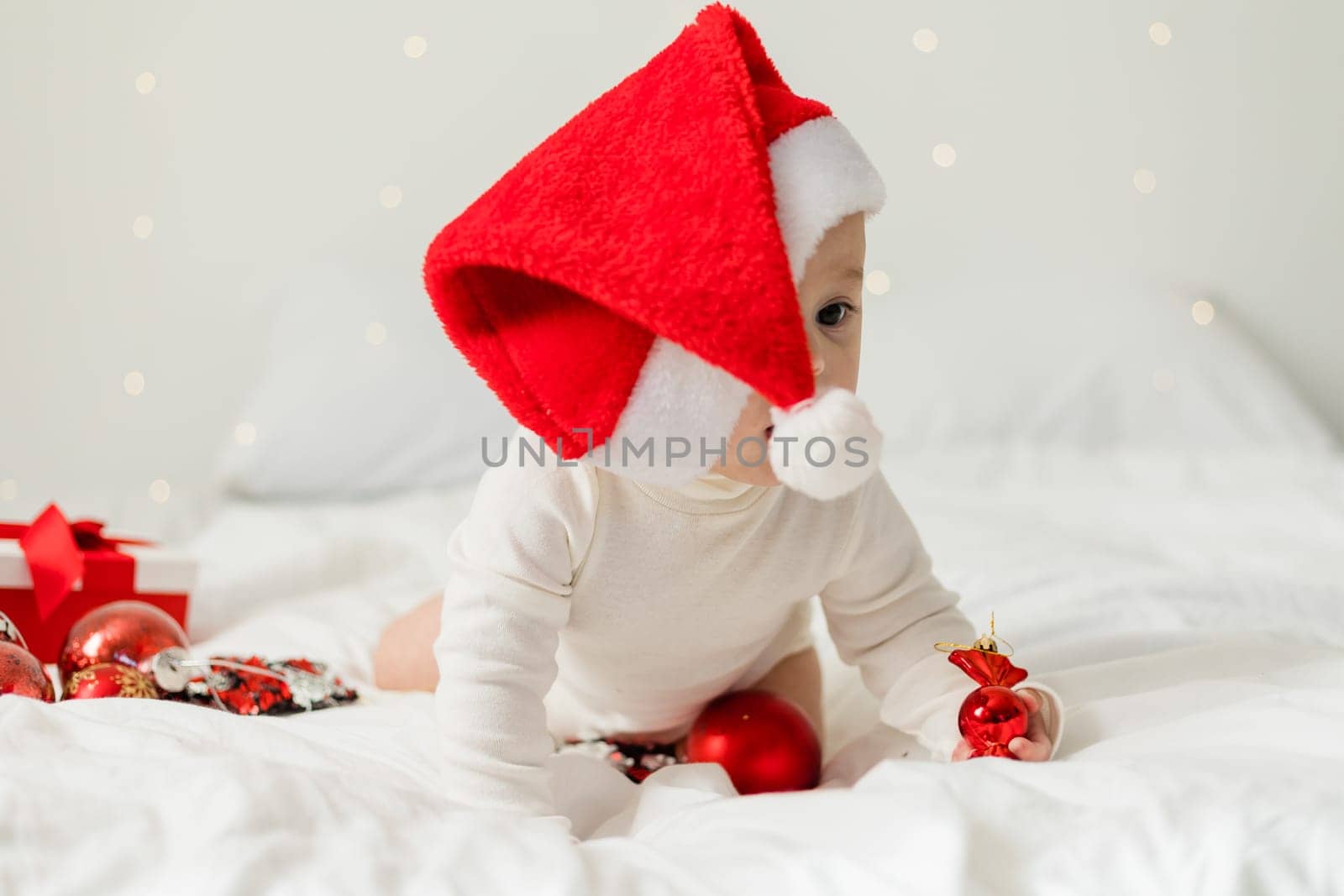 Christmas toddler in Santa hat sitting on bed with gift box and festive baubles.