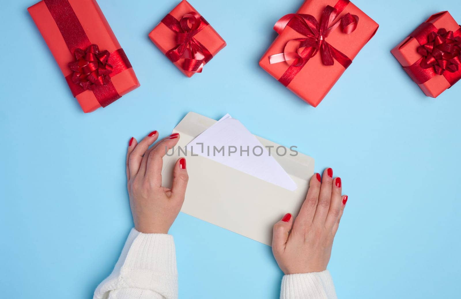 Female hands holding a white paper envelope on a blue background, around a box with gifts, top view
