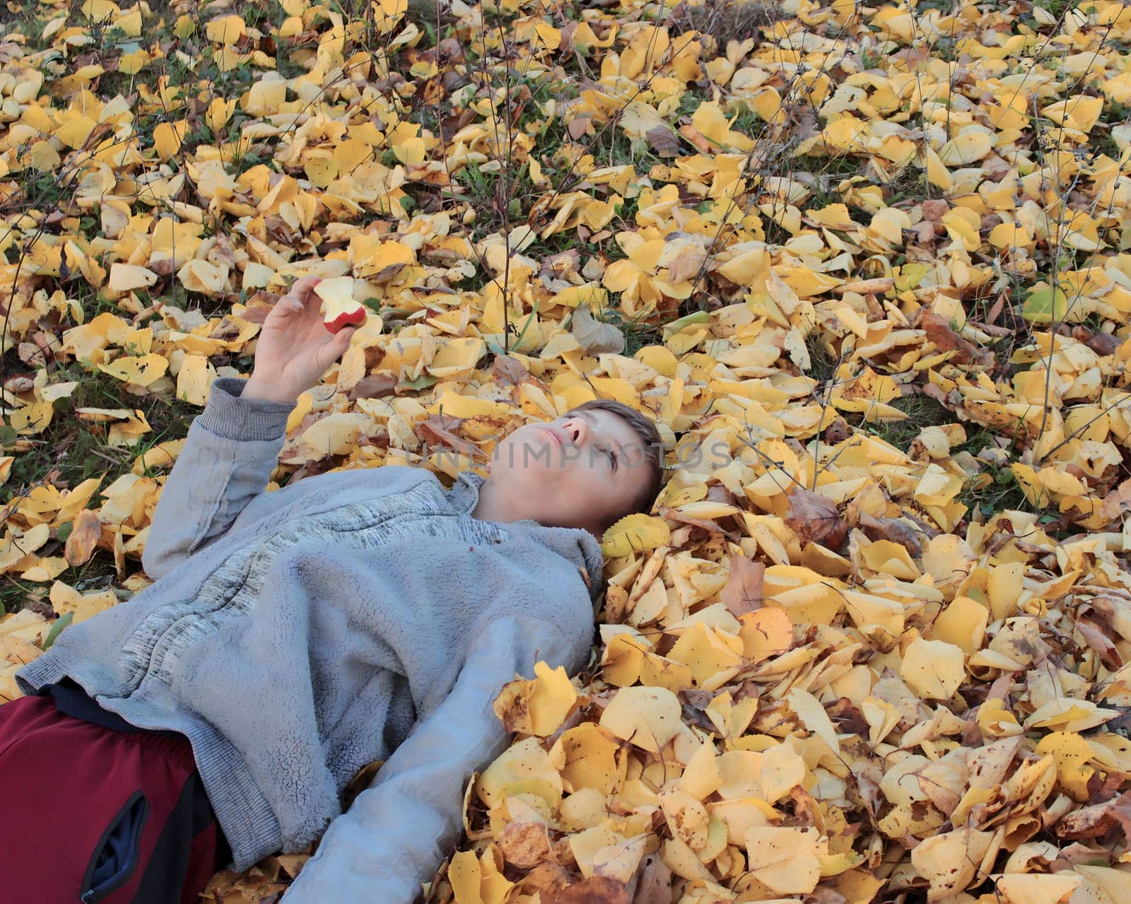 A boy lies on an autumn yellow leaf and eats an apple .