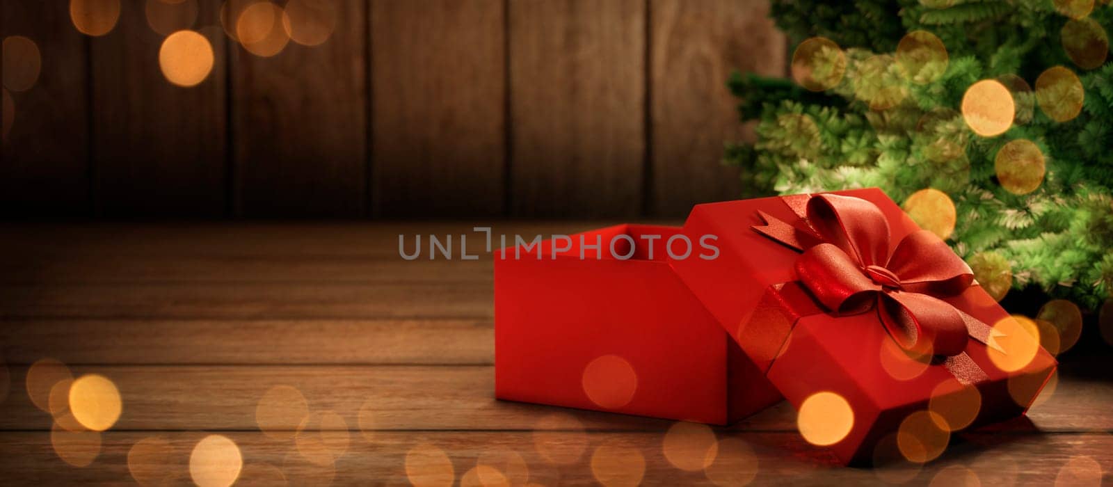 Red christmas present tied with ribbon under christmas tree with lights. Cozy magic xmas and new year holiday scene. Open gift box on rustic wooden background with copyspace.
