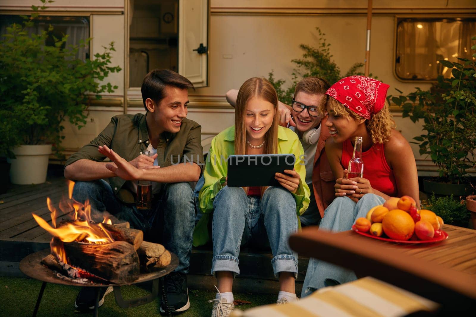 Happy friends spending evening nearby bonfire and campsite. Four millennial people watching video using mobile tablet smiling and laughing. Carefree weekend time on nature concept