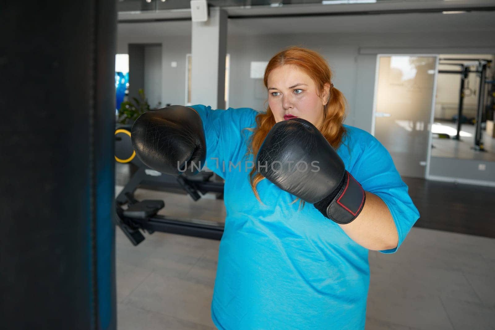 Concentrated fat woman using boxing gloves and bag doing punching exercises in gym. Sports motivation and healthy lifestyle