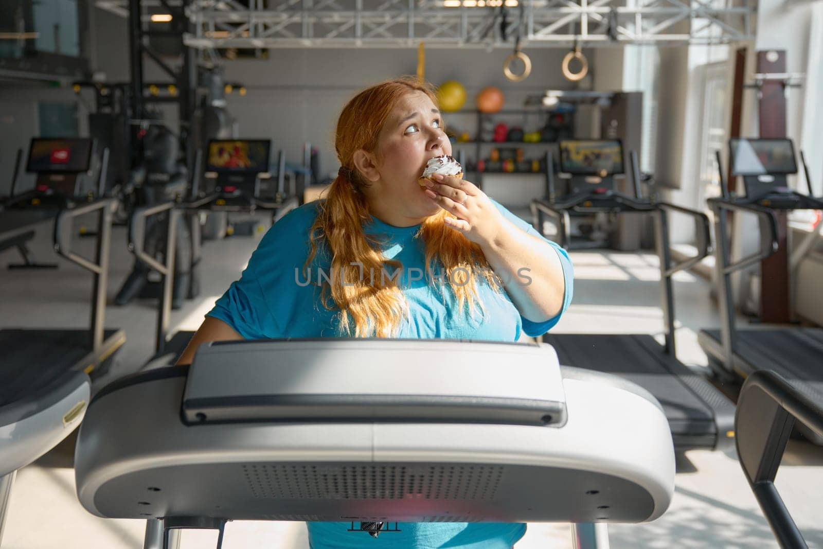 Front view on funny obese woman eating cake while training on treadmill at sport gym. Unhealthy eating disorder and fitness workout struggle